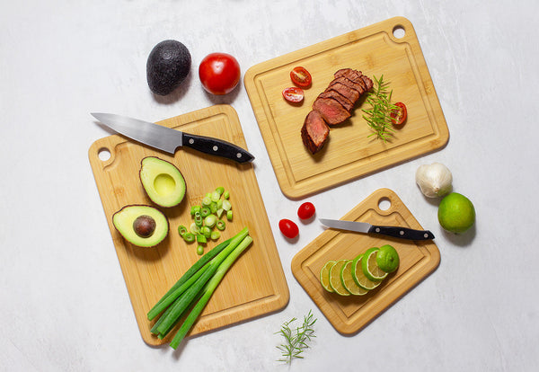Sliced steak on a wooden cutting board with a knife. Surrounded by rosemary, a bowl of garlic, salt, peppercorns, and lemons on a white marble surface. A patterned cloth is nearby.