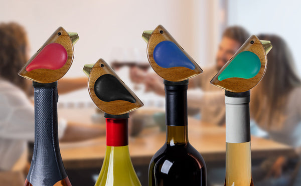 Colorful bird-shaped bottle stoppers on wine bottles with two people blurred in the background, raising glasses for a toast.