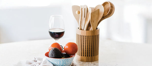 A bamboo utensil holder with wooden kitchen tools next to a glass of red wine and a bowl of tomatoes and avocados on a countertop.