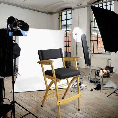 In a photography studio setup, a 32" Deluxe Bamboo Director's Chair by Totally Bamboo is positioned in front of a white backdrop. Surrounding the chair is an arrangement of lighting equipment and reflectors, while expansive windows behind allow natural light to stream in, crafting the perfect setting for capturing Hollywood-style photographs.