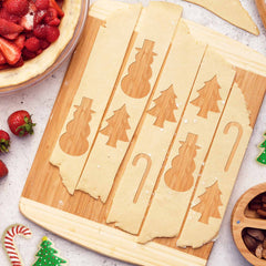 The Kauai Cutting Board by Totally Bamboo showcases strips of dough featuring cutouts of snowmen, Christmas trees, and candy canes. Nearby, a pie topped with strawberries and sugar is accompanied by whole strawberries and sugar cookies decorated in the shapes of candy canes and trees.