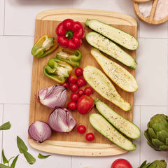 The Kona Groove Carving Board by Totally Bamboo showcases sliced zucchini, a red bell pepper, a green bell pepper, halved red onions, cherry tomatoes, and a whole tomato. These ingredients are arranged on the 18" x 12-1/2" bamboo board, surrounded by a white tiled surface with a few leaves and an artichoke nearby for impeccable kitchen prep.