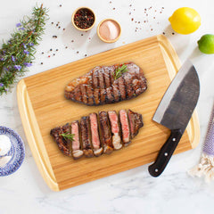 Two grilled steaks sit on the Totally Bamboo Kona Groove Carving Board, 18" x 12-1/2"; one whole and one sliced, garnished with rosemary. A large knife is positioned beside them. Encircling the board are small bowls of seasoning, a lemon, and assorted herbs, enhancing the elegance of this carving board for effortless kitchen prep.