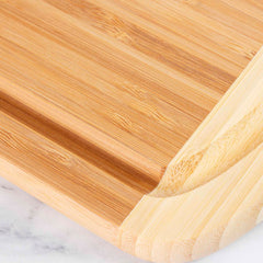 A close-up of a bamboo cutting board, similar to the Totally Bamboo Kona Groove Carving Board, measuring 18" x 12-1/2", with a smooth surface and rounded edges, resting on a light marble countertop. The grain and texture of the wood are clearly visible, making it ideal for kitchen prep tasks.