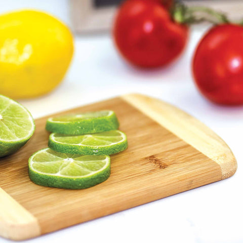 A sliced lime rests on a Totally Bamboo 2-Tone Small Cutting Board, measuring 8" x 5-3/4", with a whole lime and tomatoes in the background. A lemon is also visible, all arranged on a light surface that accentuates the rich texture of caramelized bamboo.