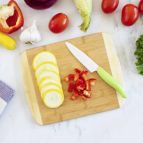 A Totally Bamboo 2-Tone Cutting Board, measuring 11" x 8-3/4", holds sliced yellow squash and diced red bell pepper beside a green-handled knife. Surrounding the kitchen scene are fresh vegetables such as tomatoes, corn, garlic, red onion, and lettuce resting on a marble surface.