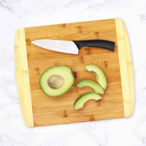 A Totally Bamboo 2-Tone Cutting Board, measuring 13-1/2" x 11-1/2", elegantly displays a ceramic knife and a sliced avocado on a pristine marble surface, perfect for any kitchen setting.