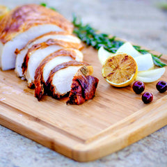 The Malibu Groove Carving Board by Totally Bamboo, measuring 18" x 12", provides a rustic and appetizing kitchen prep presentation. Sliced roasted meat is attractively arranged on the bamboo board, accompanied by fresh rosemary, halved lemon, onion slices, and dark berries.