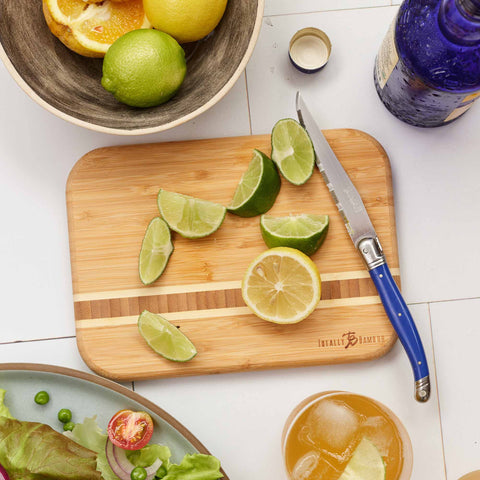 A Barbados Small Cutting Board by Totally Bamboo, measuring 9" x 6-1/2", graces the table with sliced limes and a lemon, its vibrant scene completed by a blue-handled knife resting alongside. Nearby, a bowl brims with more citrus fruits from the Caribbean Collection, while a blue bottle complements the salad and glass of refreshing yellow drink.