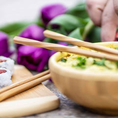 A close-up of a meal featuring a wooden bowl with noodles and vegetables, accompanied by Totally Bamboo's "Twist" Reusable Bamboo Chopsticks resting on its rim. To the left, sushi pieces are arranged on a wooden board. In the background, purple tulips appear slightly blurred.