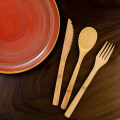 A ceramic plate with a red and orange gradient is placed next to the 3-Piece Reusable Bamboo Flatware Set by Totally Bamboo, featuring a knife, spoon, and fork, on a dark wooden surface.