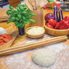 On a countertop, fresh pizza dough rests on a floured surface, accompanied by the Totally Bamboo Tapered Rolling Pin, 20-1/2" x 1-3/4". Nearby, a basil plant and bowls containing shredded cheese, pepperoni slices, tomatoes, red onion pieces, red peppers, and eggplants surround the scene. Kitchen utensils are neatly arranged in a holder close by.