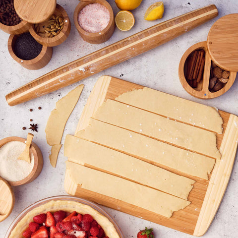 A Totally Bamboo cutting board made from Moso bamboo features strips of dough, accompanied by a Tapered Rolling Pin (20-1/2" x 1-3/4") and bowls filled with various spices and ingredients. Below, you can see a partially visible pie with fruit filling. The scene is neatly arranged on a light countertop.