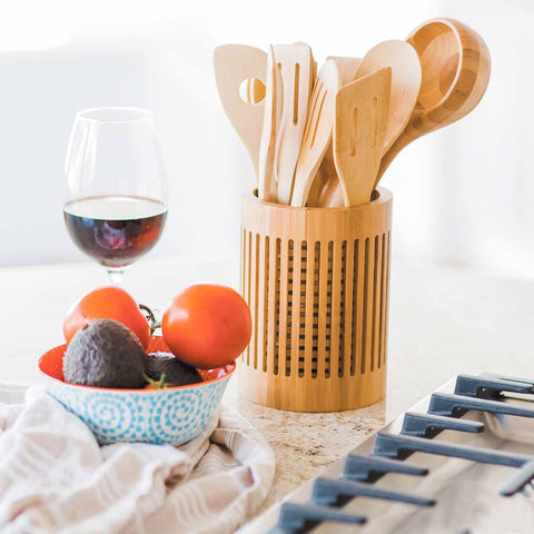 A Totally Bamboo Lattice Kitchen Utensil Holder with wooden cooking tools sits on a counter beside a glass of red wine. Nearby, a blue bowl containing avocados and tomatoes rests partly on a white dish towel, while a stovetop is visible in the foreground.