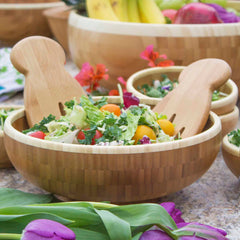 A Moso bamboo salad bowl filled with mixed greens, cherry tomatoes, and colorful vegetables is accompanied by the elegant Totally Bamboo Salad Hands Salad Serving Set. This delightful arrangement is set against a rustic stone surface, surrounded by fresh fruits and other salad bowls.