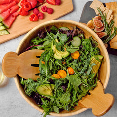 A wooden bowl filled with fresh green salad, topped with sliced avocado and cherry tomatoes, is ready to be served with the Salad Hands Salad Serving Set by Totally Bamboo. In the background, a cutting board showcases strawberries and watermelon next to a plate of crackers and figs.