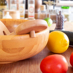 A wooden mortar and pestle rest on a kitchen counter alongside a lemon, lime, tomato, and cucumber. Nearby, the stylish Salad Hands Salad Serving Set from Totally Bamboo enhances the scene. In the background, spices and condiments are slightly blurred, adding depth to the culinary tableau.