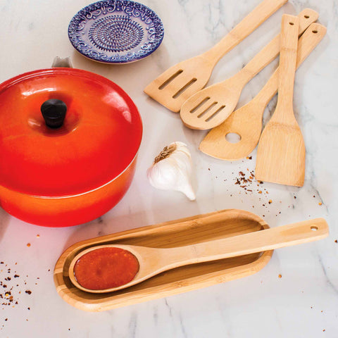 A red pot, renowned for its non-stick cookware safety, is placed on a marble surface next to the 5-Piece Bamboo Cooking Utensil Set by Totally Bamboo, a garlic bulb, and a blue patterned dish. A spoon lies on a tray of tomato sauce with crushed chili flakes sprinkled nearby.