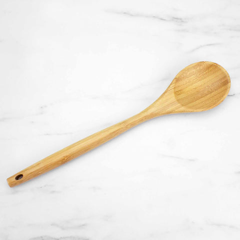 A Totally Bamboo "Lambootensil" Cooking Spoon, 14", sits on a white marble surface. This bamboo cooking spoon includes an oval-shaped head and a long handle with a small hole at the end for convenient hanging, making it ideal for use with nonstick safe cookware.