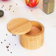 A Totally Bamboo Salt Cellar with a swivel lid is partially open, showcasing pink Himalayan salt inside its 6-ounce capacity. Nearby, black peppercorns are scattered across a white surface, while an apple and a spice jar remain in the background, ideal companions for this elegant seasoning storage solution.