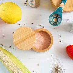 A Totally Bamboo salt cellar with a swivel lid, holding 6 ounces of pink salt, is surrounded by a lemon, uncooked corn, a teal wooden spoon, black peppercorns, and a red bell pepper on a white marble surface.