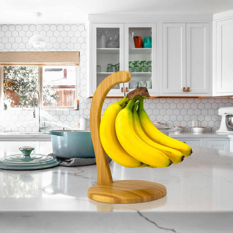 A bunch of bananas hangs on a Totally Bamboo banana holder with a stainless steel hook, placed on a white kitchen island. The kitchen features white cabinets, a hexagonal tile backsplash, and a window. A teal pot and stand mixer glisten in the background under the soft light filtering through.