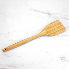 A "Lambootensil" Spatula by Totally Bamboo, measuring 14 inches and made from Moso bamboo, featuring a wide, flat head and a long handle, elegantly rests on a white marble surface. Ideal for use with nonstick cookware.