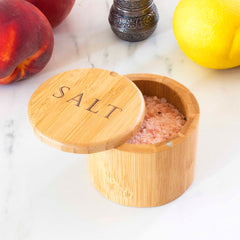 A Totally Bamboo "Salt" Engraved Salt Cellar with a swiveling lid is open, showcasing pink salt inside. With a 6-oz. capacity, its magnetic lock ensures secure closure. It rests on a marble surface beside a peach, a lemon, and a small decorative pepper grinder.