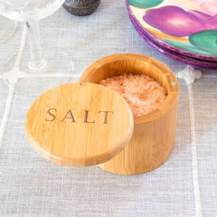 A "Salt" Engraved Salt Cellar by Totally Bamboo, featuring a magnetic swiveling lid, sits open on a gray tablecloth. Its 6-ounce capacity is filled with pink Himalayan salt crystals. In the background, you can see part of a colorful plate and wine glass.