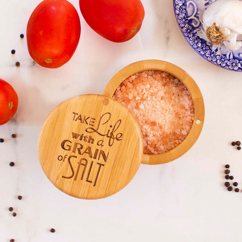 On a white marble surface, an open "Grain of Salt" Engraved Salt Cellar by Totally Bamboo reveals pink Himalayan salt. Its swiveling lid is slightly open, surrounded by fresh tomatoes, garlic cloves, and scattered black peppercorns.