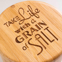 An engraved wooden surface displaying the phrase "Take Life with a Grain of Salt" in artistic, varied fonts captures the essence of the "Grain of Salt" Engraved Salt Cellar by Totally Bamboo—a charming 6-oz. capacity accessory with a swiveling lid that combines elegance and practical appeal.