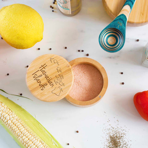 A Totally Bamboo "Hawaiian Islands" Engraved Salt Cellar, featuring a 6-oz. capacity, is open on a marble surface and filled with pink salt. Surrounding it are lemon, corn, tomato, a blue-striped wooden spoon, peppercorns, and dried herbs. The magnetic lock of the cellar ensures that your kitchen is both fresh and stylish.