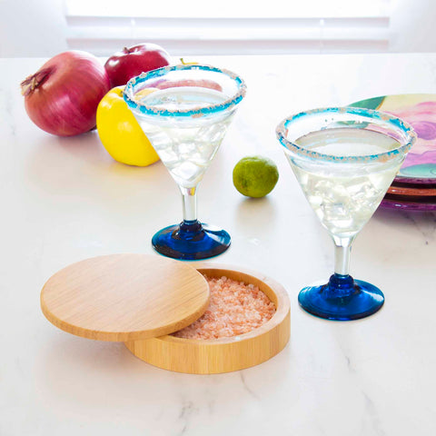 Two margarita glasses with salted rims rest on a marble countertop beside a Totally Bamboo "Barkeeper's Salt Box" Margarita Glass Rimmer filled with pink salt, alongside a lime, a lemon, and two red onions. In the background, a stack of colorful plates adds a vibrant touch to the scene.