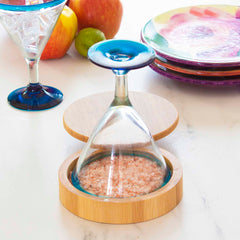 A Barkeeper's Salt Box Margarita Glass Rimmer from Totally Bamboo, featuring a blue-stemmed wine glass inverted and its bowl filled with pink salt, rests in a Moso bamboo holder. This setup is displayed elegantly on a marble surface, accented by colorful plates and fruit in the background.