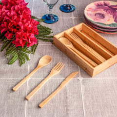 A 12-piece reusable bamboo flatware set by Totally Bamboo, made from durable Moso bamboo, features a spoon, fork, and knife on a beige tablecloth. Accompanying the scene is a wooden organizer and a bright bouquet of red flowers accented with ferns. Two wine glasses and a stack of floral plates complete the sophisticated dining arrangement.