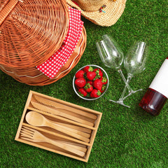 The picnic setup on grass includes a wicker basket, a 12-piece reusable bamboo flatware set from Totally Bamboo, fresh strawberries in a bowl, two wine glasses, and a bottle of wine with a straw hat resting nearby.