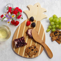 The Pineapple Serving Board with Spreader Knife Gift Set by Totally Bamboo offers a tropical style ensemble for any gathering. The board elegantly showcases cheese, fruit spreads, crackers, raspberries, blackberries, and grapes. Beside it is the bamboo spreader knife included in the set, complemented by a glass of white wine and a small flower bouquet on a white surface.