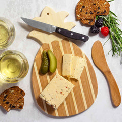 The Pineapple Serving Board with Spreader Knife Gift Set by Totally Bamboo, showcasing a tropical style, elegantly holds slices of cheese and pickles along with a bamboo spreader knife. On the light surface nearby, cranberry crisps, berries, sprigs of rosemary, and two glasses of white wine complete the scene.