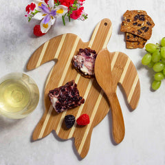 The Sea Turtle Serving Board with Spreader Knife Gift Set from Totally Bamboo showcases two berry cheese slices and a bamboo spreader knife, surrounded by assorted berries. Nearby, you'll find crackers, a glass of white wine, grapes, and a vibrant floral arrangement on a light surface.