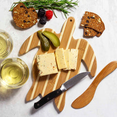 Cheese slices and a pickle are elegantly presented on the Totally Bamboo Sea Turtle Serving Board. Accompanied by rye crisps, berries, and rosemary, this set includes a bamboo spreader knife from the gift collection. Nearby are two glasses of white wine and a sleek black-handled knife.