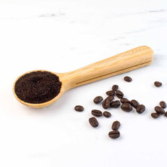 A Coffee Scoop with Built-In Bag Clip by Totally Bamboo cradles freshly ground coffee on a white surface, surrounded by a few scattered beans.