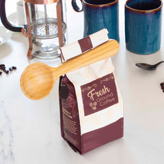 A bag of freshly ground coffee, secured with a Totally Bamboo Coffee Scoop with Built-In Bag Clip, sits on a white countertop. In the background, a French press, blue mugs, and scattered coffee beans evoke a cozy morning ritual.