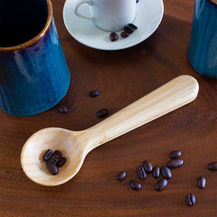 A Totally Bamboo Coffee Scoop with Built-In Bag Clip and beans rests on a wooden table. Nearby, scattered coffee beans surround a blue ceramic mug, another blue mug on its side, and a white espresso cup on a saucer.