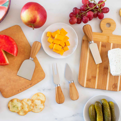 A flat lay image of a cheese platter setup features the Totally Bamboo 4-Piece Cheese Knife Set with stainless steel blades and bamboo handles, arranged alongside assorted cheeses on boards, sliced red apple, red grapes, sliced baguette, pickles, and crackers on a white surface.