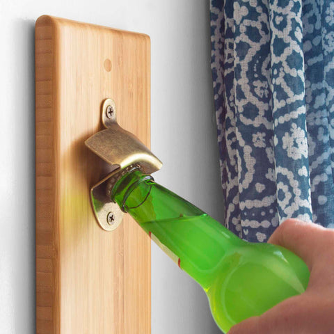 A person uses the Totally Bamboo Wall-Mounted Bottle Opener with Magnetic Cap Catcher on a wooden board to open a green glass bottle, with a blue patterned curtain visible in the background.