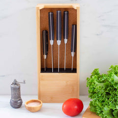 A set of five sleek kitchen knives with black handles are neatly displayed in the Totally Bamboo Universal Knife Caddy. In front of the caddy, you can see a pepper grinder, a small bowl of salt, a ripe tomato, and fresh leafy greens on a pristine white surface.