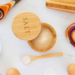 The "Salt" Engraved Little Dipper Salt Cellar with Salt Spoon by Totally Bamboo is open, showcasing coarse salt. Alongside it on a white surface lies the small bamboo serving spoon heaped with salt, accompanied by two eggs and vibrant wooden utensils.