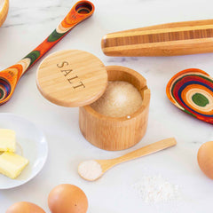 A kitchen scene showcases the "Salt" Engraved Little Dipper Salt Cellar with a salt spoon from Totally Bamboo, accompanied by a magnetic-lock salt cellar, colorful measuring spoons, a rolling pin, eggs, and butter on a plate. Everything is neatly arranged on a pristine white marble countertop.