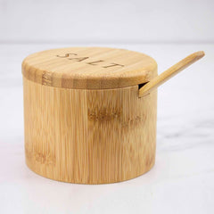 The "Salt" Engraved Little Dipper Salt Cellar with Salt Spoon by Totally Bamboo is a round, wooden salt cellar featuring "SALT" etched on the lid and includes a bamboo serving spoon resting in a notch. Enhanced with a magnetic lock, it is elegantly displayed against a smooth, white background.