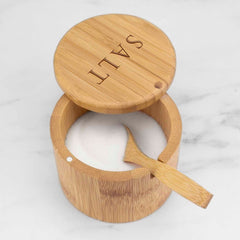 The "Salt" Engraved Little Dipper Salt Cellar with Salt Spoon by Totally Bamboo subtly displays its charm as it rests slightly open on a white marble surface. Inside, a small bamboo serving spoon awaits its use, accentuating the elegance of the piece enhanced by a gentle magnetic lock.